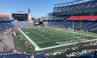 Wide view of a football stadium with blue seating, green field, and yellow goalposts; players and fans on the sidelines under a clear blue sky.