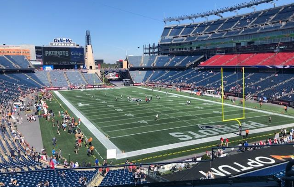 Wide view of a football stadium with blue seating, green field, and yellow goalposts; players and fans on the sidelines under a clear blue sky.