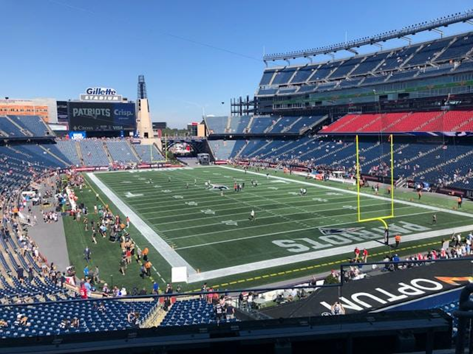 Wide view of a football stadium with blue seating, green field, and yellow goalposts; players and fans on the sidelines under a clear blue sky.